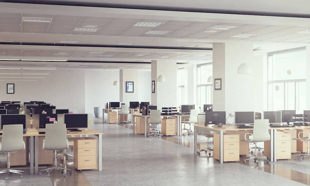 Spacious open-plan office with rows of desks, chairs, and computers, illuminated by natural light through large windows.