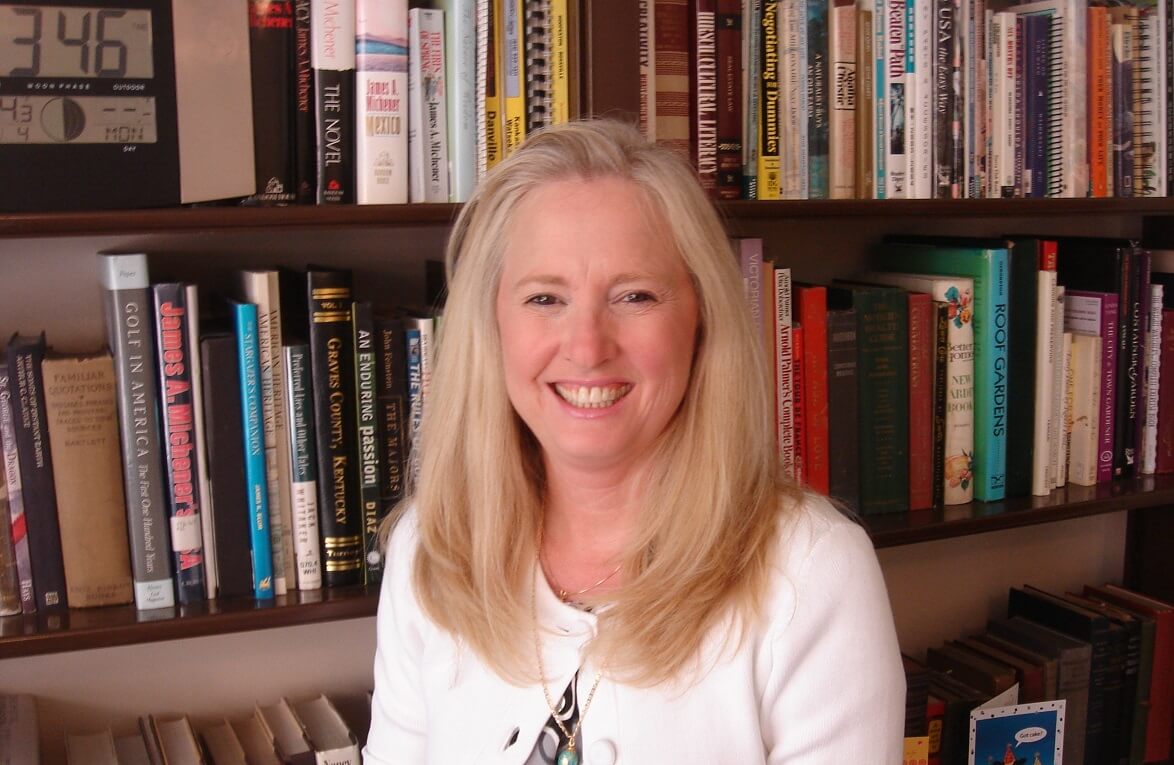 Smiling woman with long blonde hair wearing a white top, sitting in front of a bookshelf filled with various books.