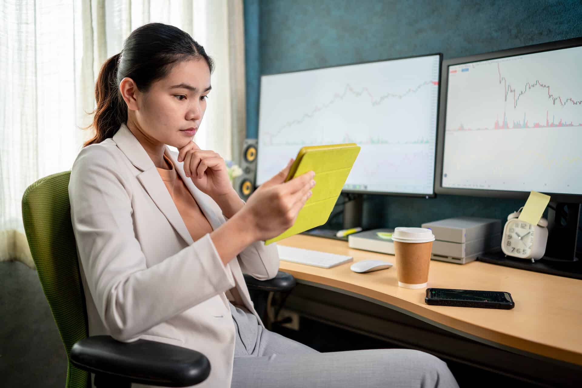 Person in an office, reading a tablet, with financial charts on dual monitors and a coffee cup on the desk.