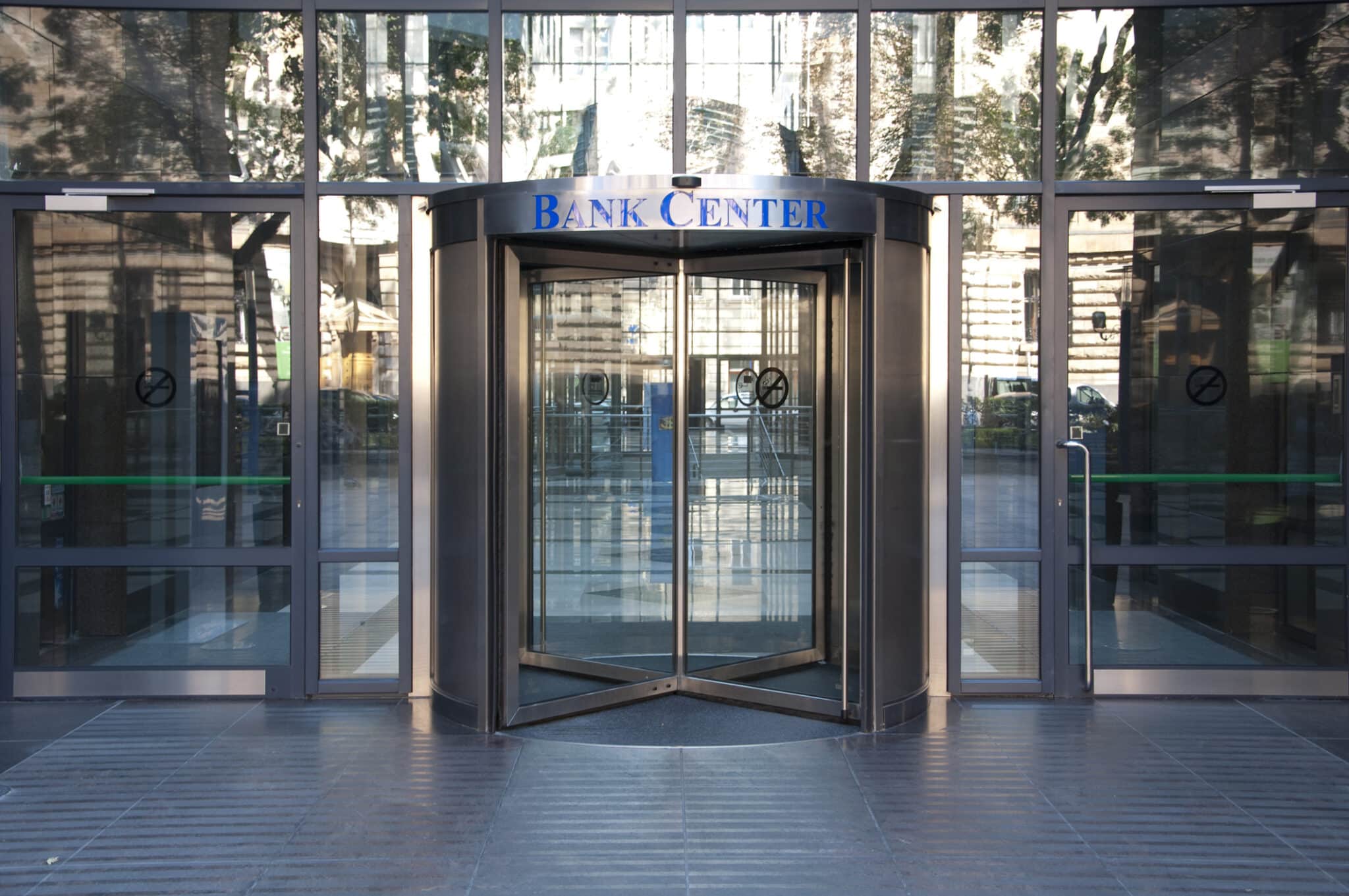 Glass revolving door entrance to a building with "Bank Center" sign above, reflecting the urban surroundings.