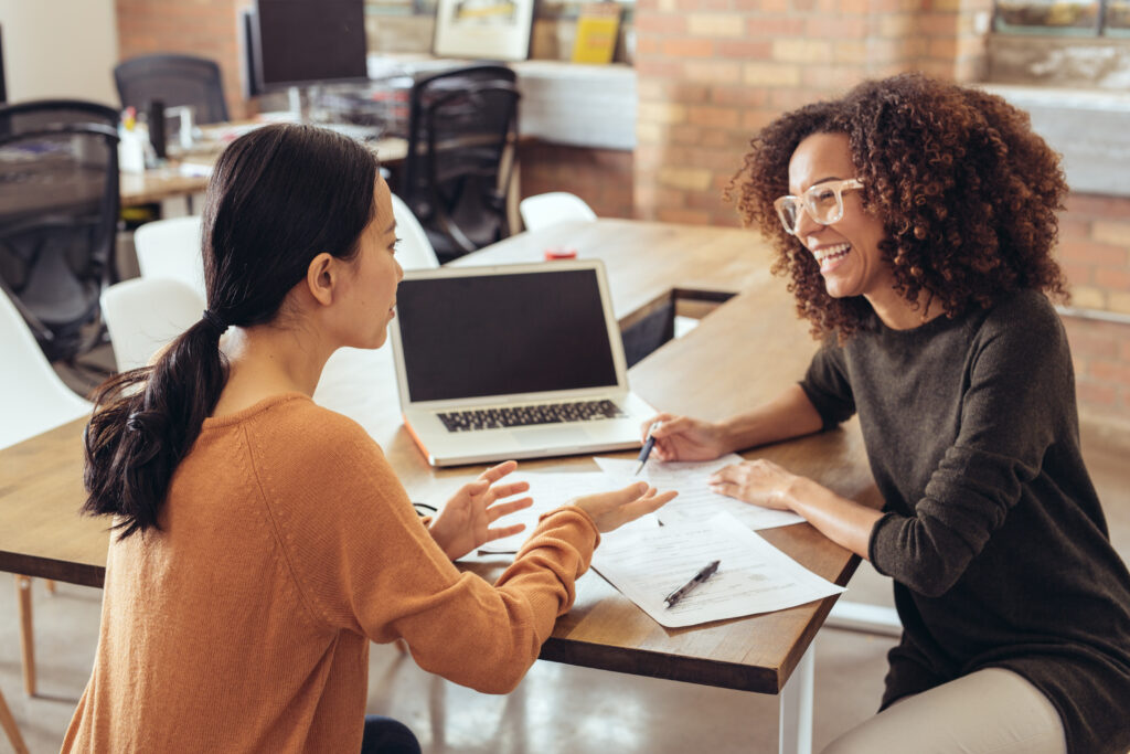 Two women working in a modern office with a laptop on the table between them,