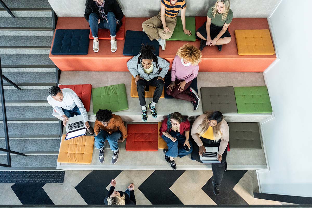 A diverse group of people sitting on tiered seating with laptops and notebooks, viewed from above.