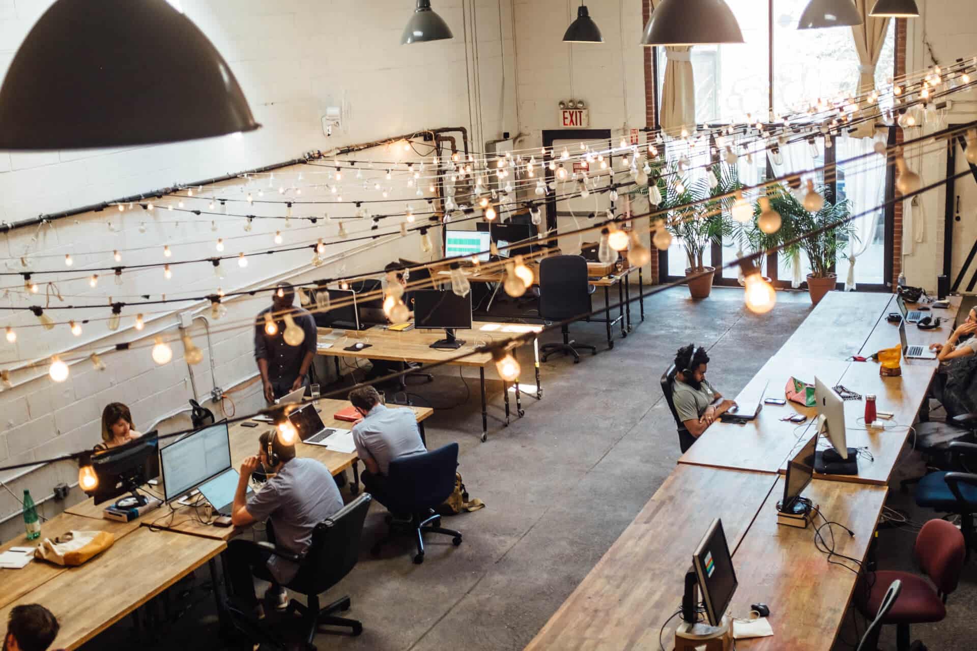 Open office space with people working at long wooden tables, decorated with hanging string lights and large lamps.